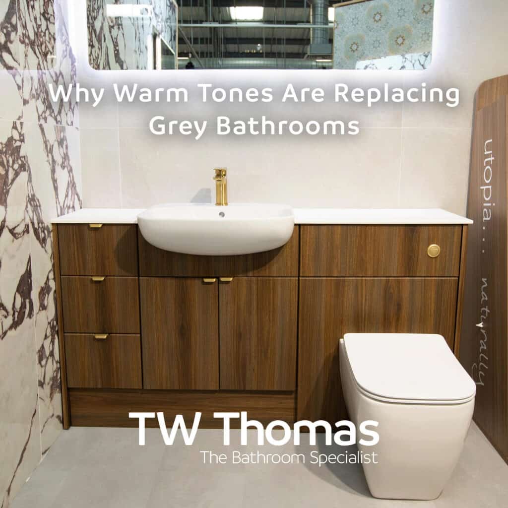 A walnut fitted furniture run with a white basin and brushed brass tap against veined marble-effect tiles, captioned Why Warm Tones Are Replacing Grey Bathrooms