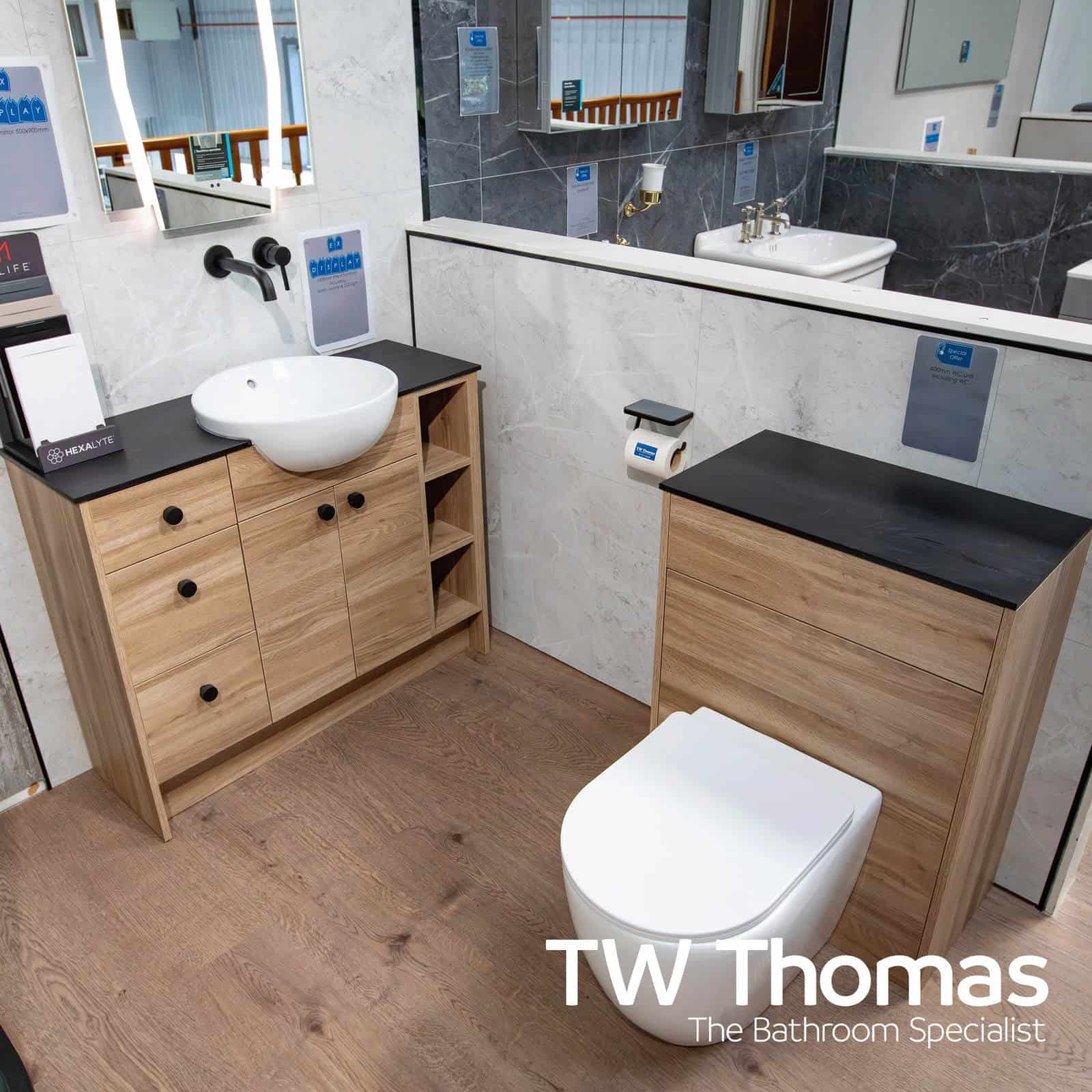 An oak-effect open vanity unit with a black worktop, round countertop basin and matt black wall taps, beside a back-to-wall WC in a showroom display