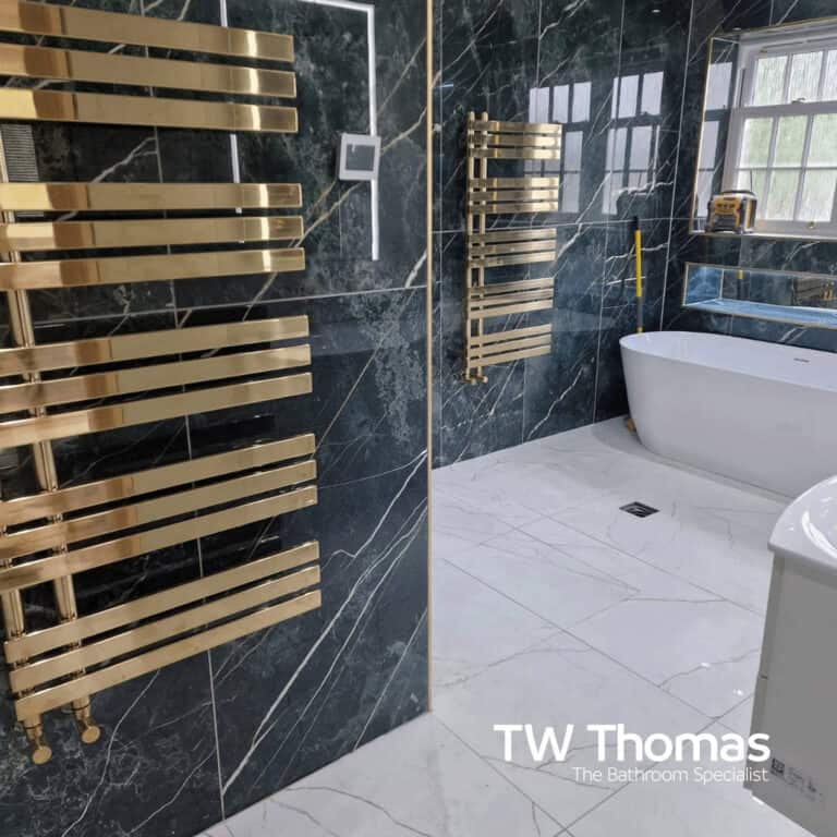 A dark marble-effect wet room with two brushed gold ladder towel radiators and a white freestanding bath on light marble floor tiles