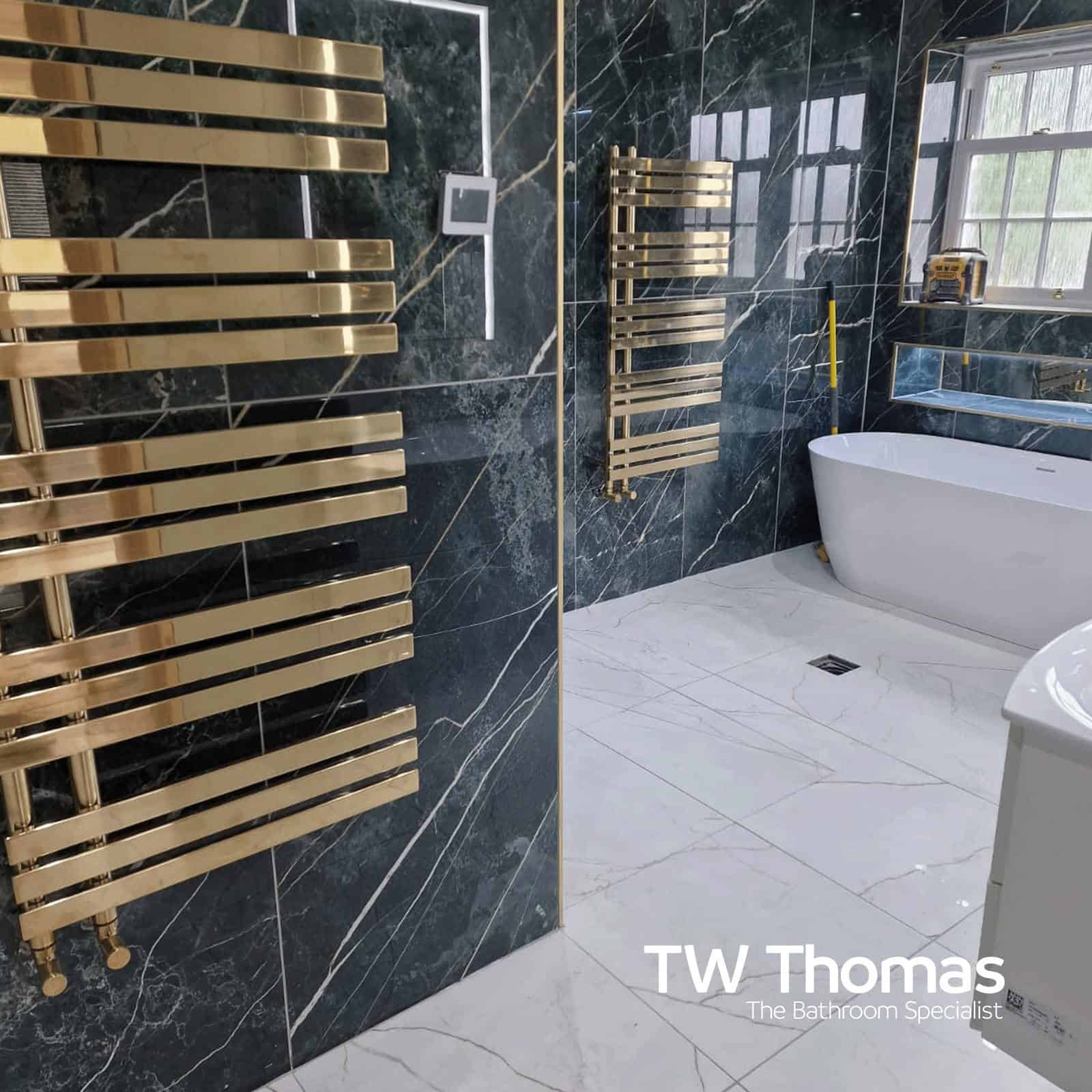 A dark marble-effect wet room with two brushed gold ladder towel radiators and a white freestanding bath on light marble floor tiles