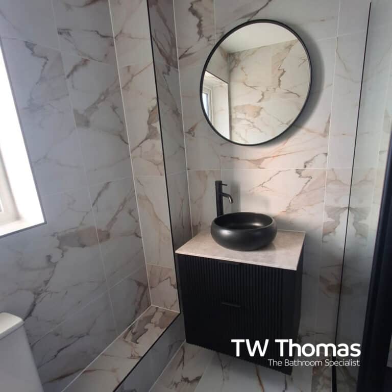 A narrow shower room in marble-effect tiles with a black ribbed wall-hung vanity, black round countertop basin and a round black-framed mirror