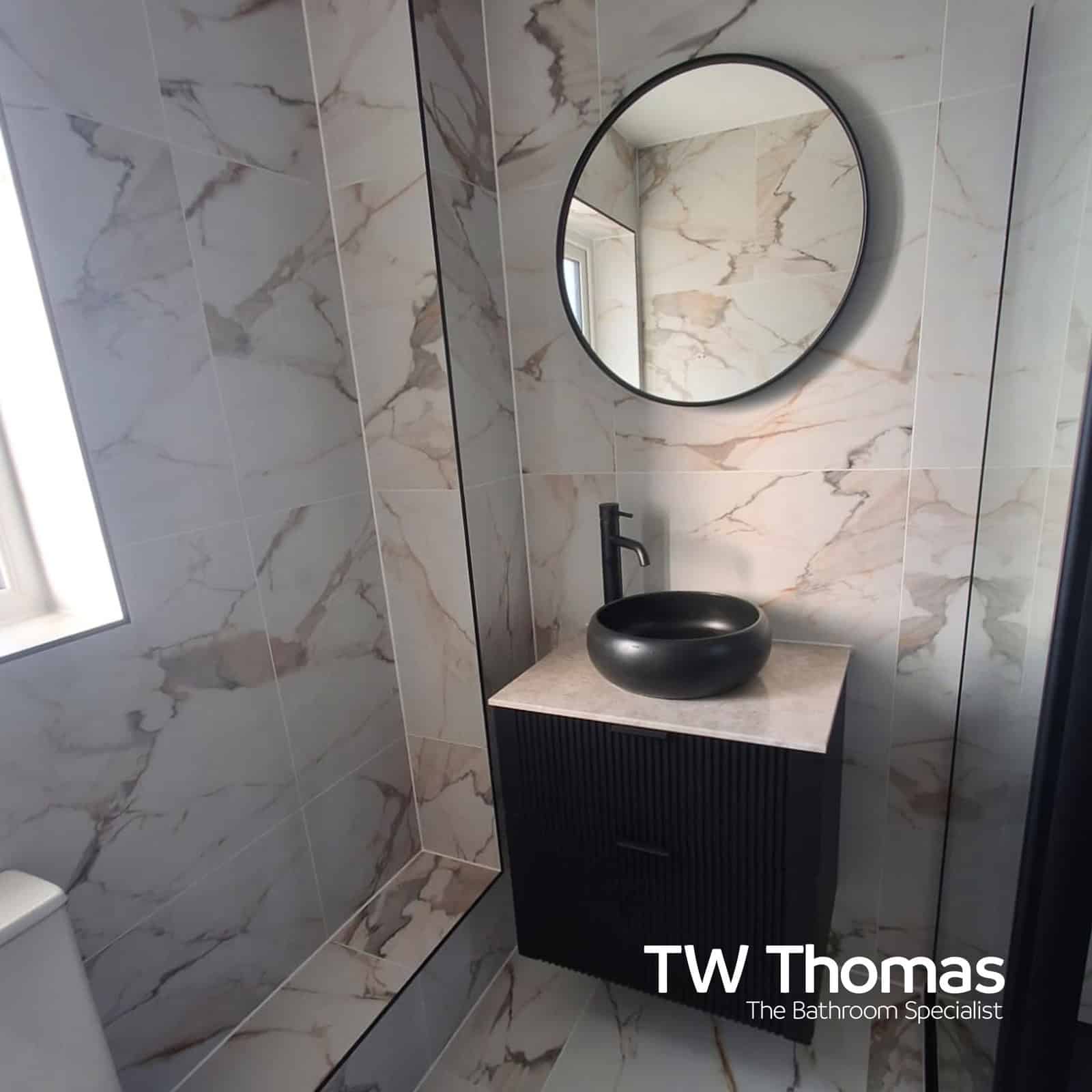 A narrow shower room in marble-effect tiles with a black ribbed wall-hung vanity, black round countertop basin and a round black-framed mirror