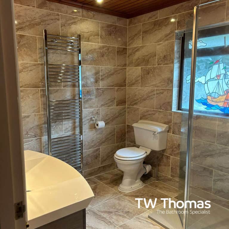A bathroom in brown marble-effect tiles with a chrome ladder towel radiator, close-coupled WC and a walk-in shower, beside a stained glass window