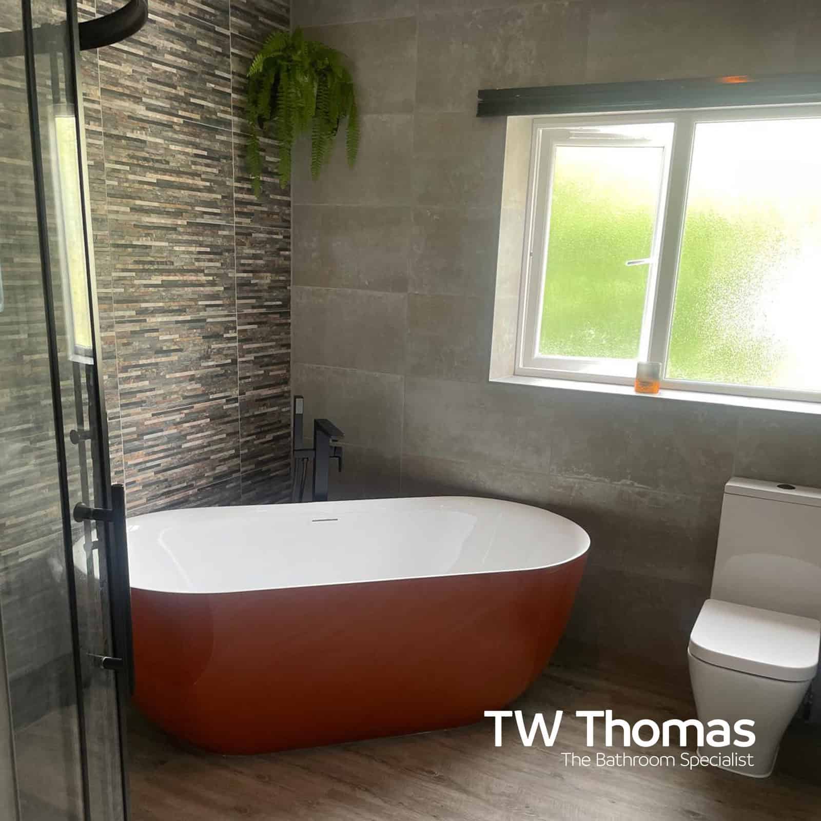 A freestanding bath with a burnt orange exterior against grey tiles and a stone strip feature wall, beside a window and a back-to-wall WC