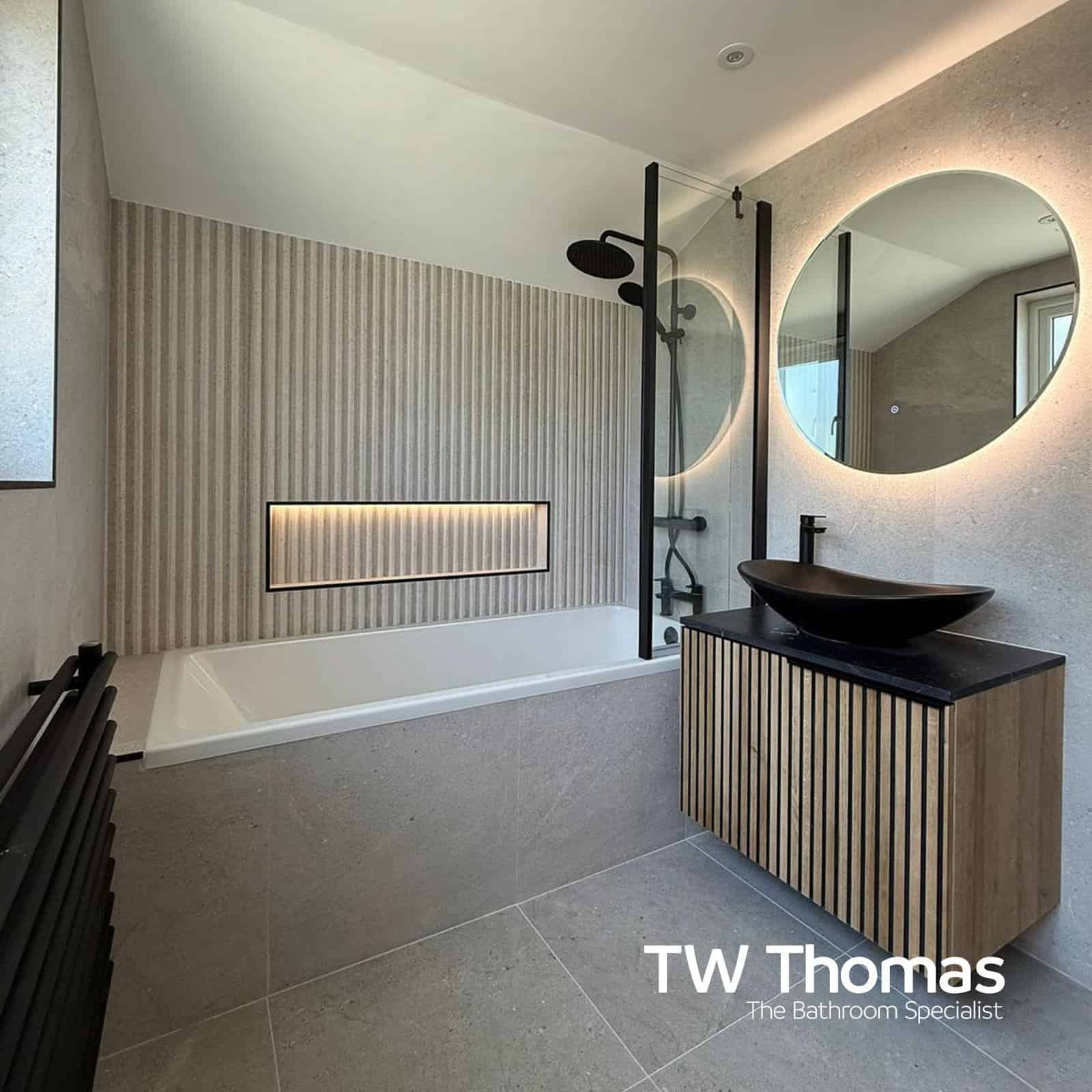 A bath with a black-framed screen and rainfall shower, two round backlit mirrors, and a ribbed wood vanity with a black oval countertop basin