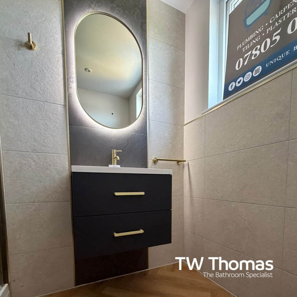 An oval backlit mirror above a dark wall-hung two-drawer vanity with brass handles and tap, against beige stone-effect tiles