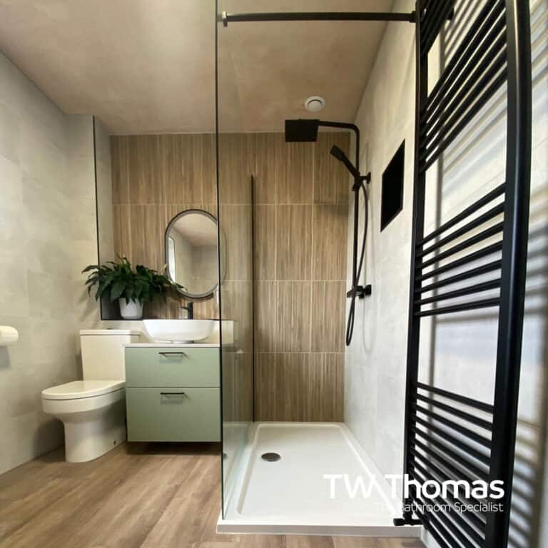 A walk-in shower with a matt black frame and rainfall head beside a sage green wall-hung vanity, oval black-framed mirror and wood-effect floor
