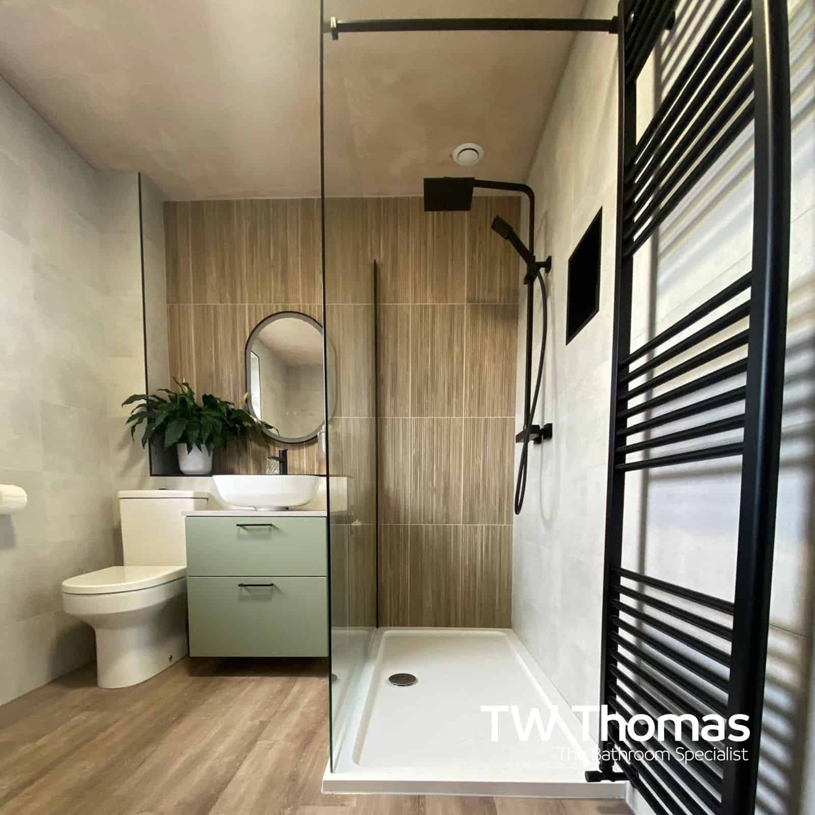 A walk-in shower with a matt black frame and rainfall head beside a sage green wall-hung vanity, oval black-framed mirror and wood-effect floor