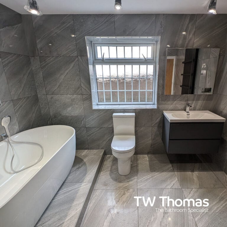 A grey marble-effect bathroom with a white freestanding bath, close-coupled WC under a window and a dark grey wall-hung vanity