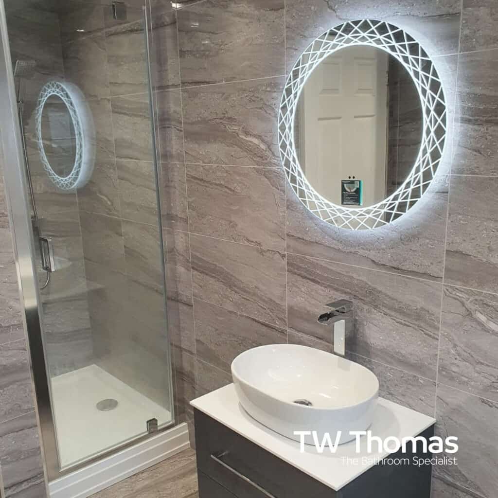 A corner shower enclosure in grey stone-effect tiles beside a grey wall-hung vanity with an oval countertop basin and a round backlit mirror