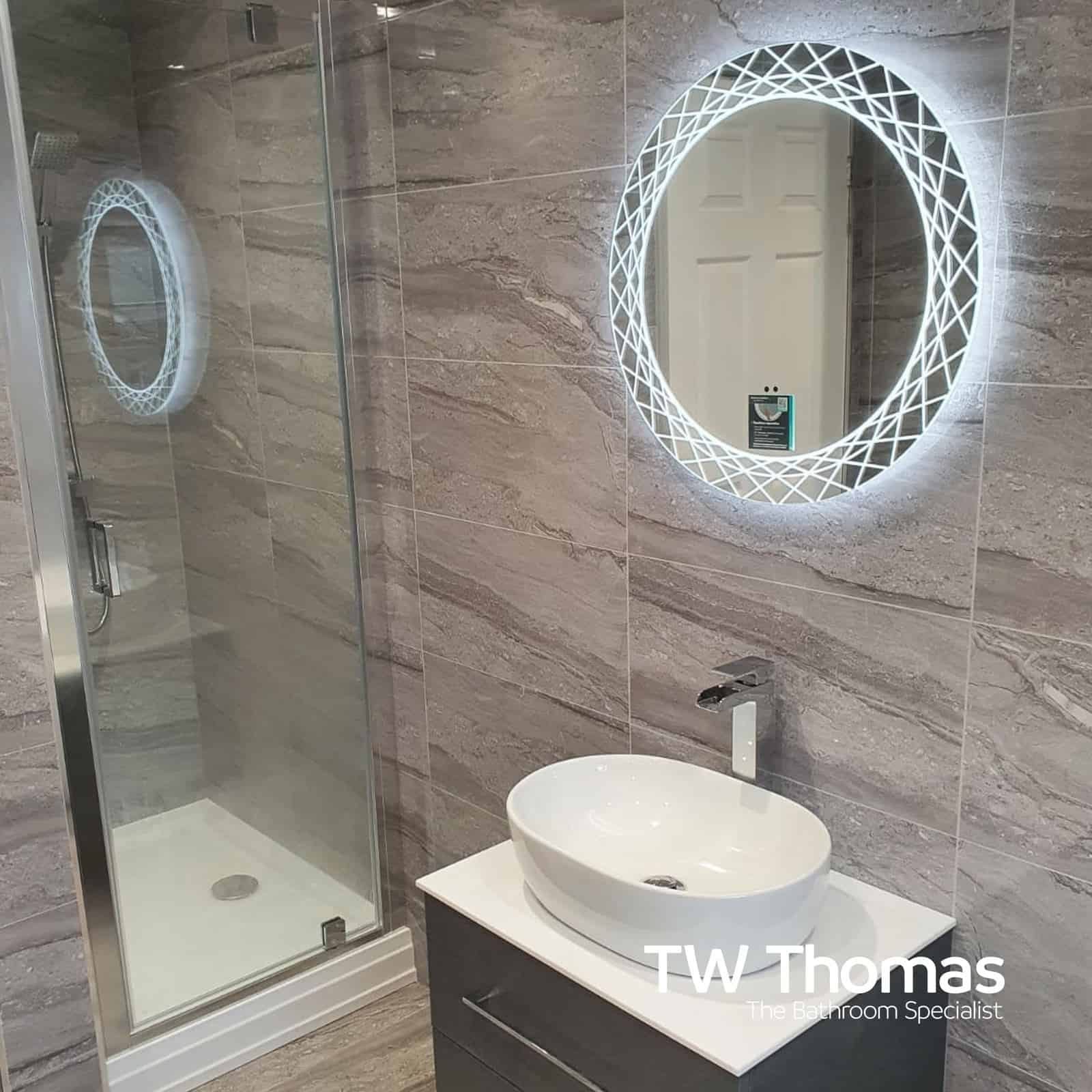 A corner shower enclosure in grey stone-effect tiles beside a grey wall-hung vanity with an oval countertop basin and a round backlit mirror