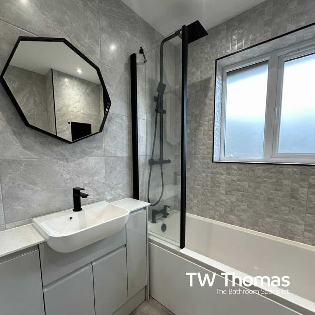 An octagonal black-framed mirror above a white basin on a light grey fitted run, beside a bath with a black screen and black shower column