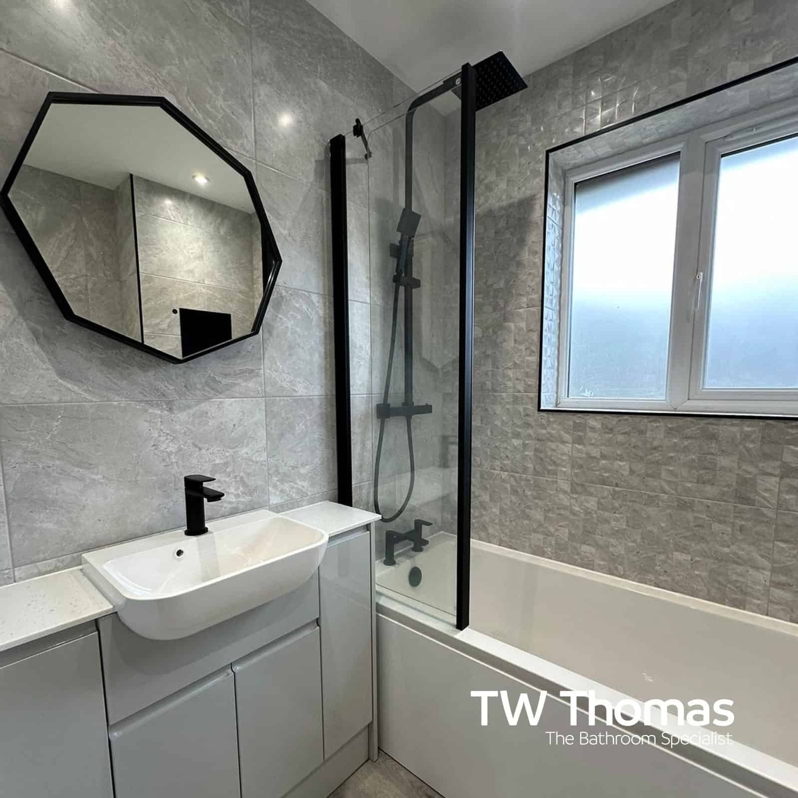 An octagonal black-framed mirror above a white basin on a light grey fitted run, beside a bath with a black screen and black shower column
