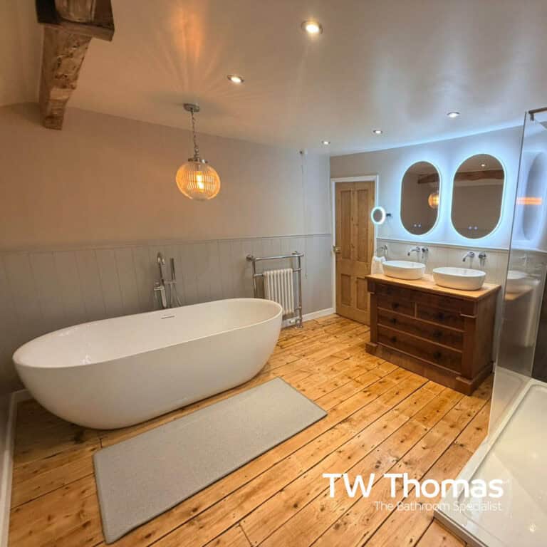 A white freestanding bath on stripped floorboards under an exposed beam, with twin backlit mirrors above a reclaimed wood double vanity