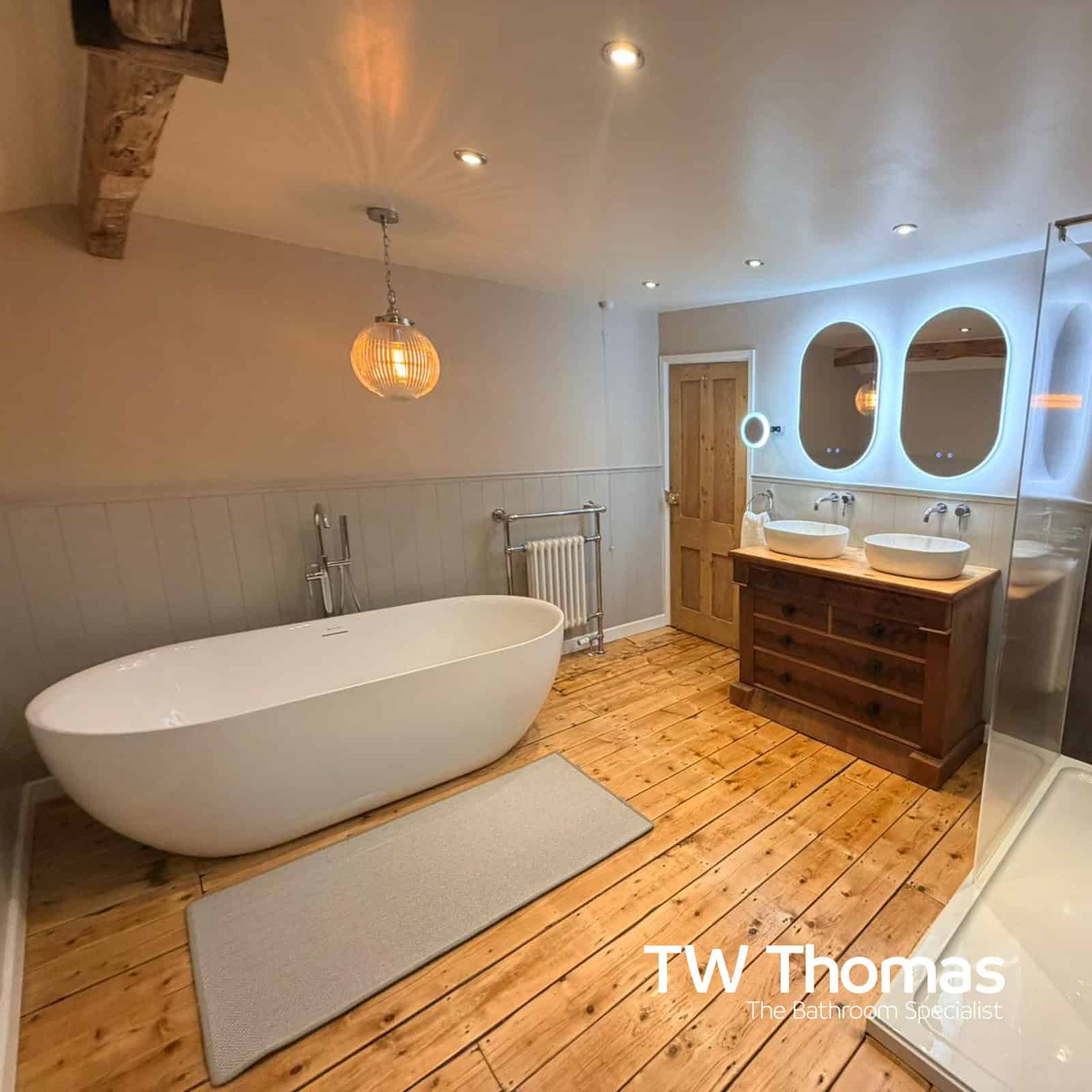 A white freestanding bath on stripped floorboards under an exposed beam, with twin backlit mirrors above a reclaimed wood double vanity