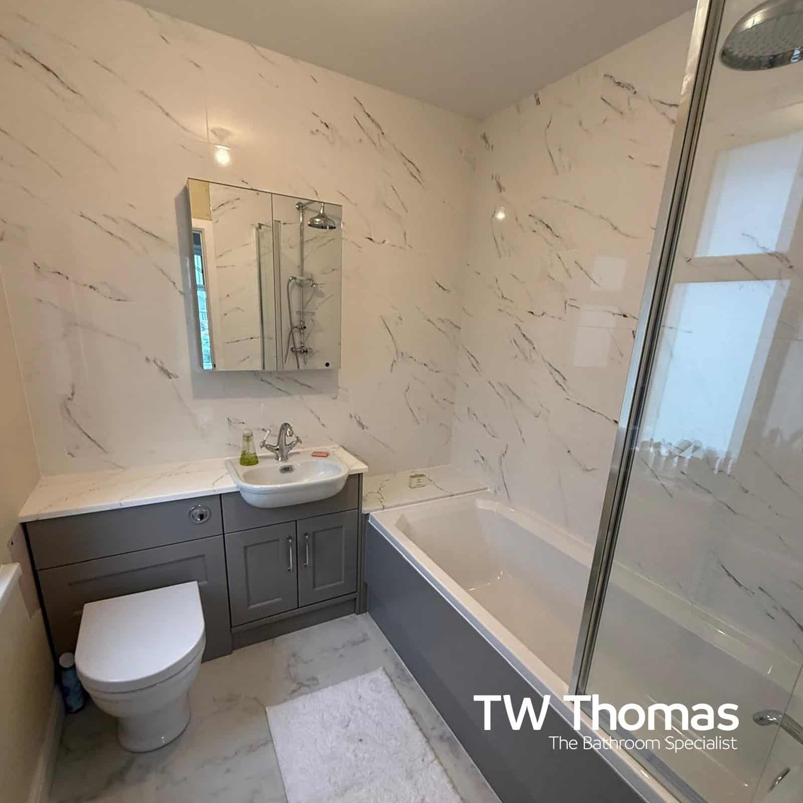 A white marble-effect bathroom with grey fitted furniture, a mirrored cabinet and a bath with a glass shower screen