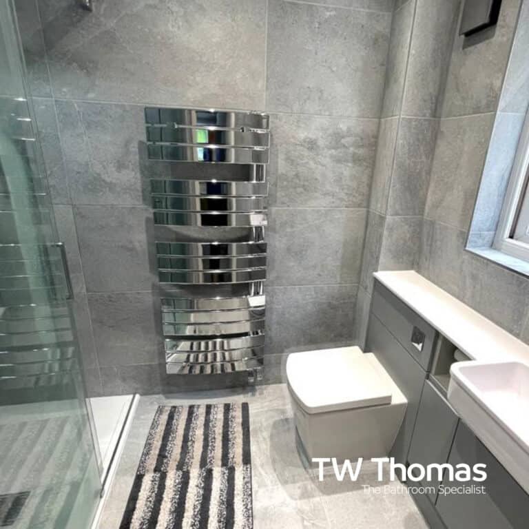 A grey tiled shower room with a large chrome designer towel radiator, a wall-hung WC and a glass shower screen