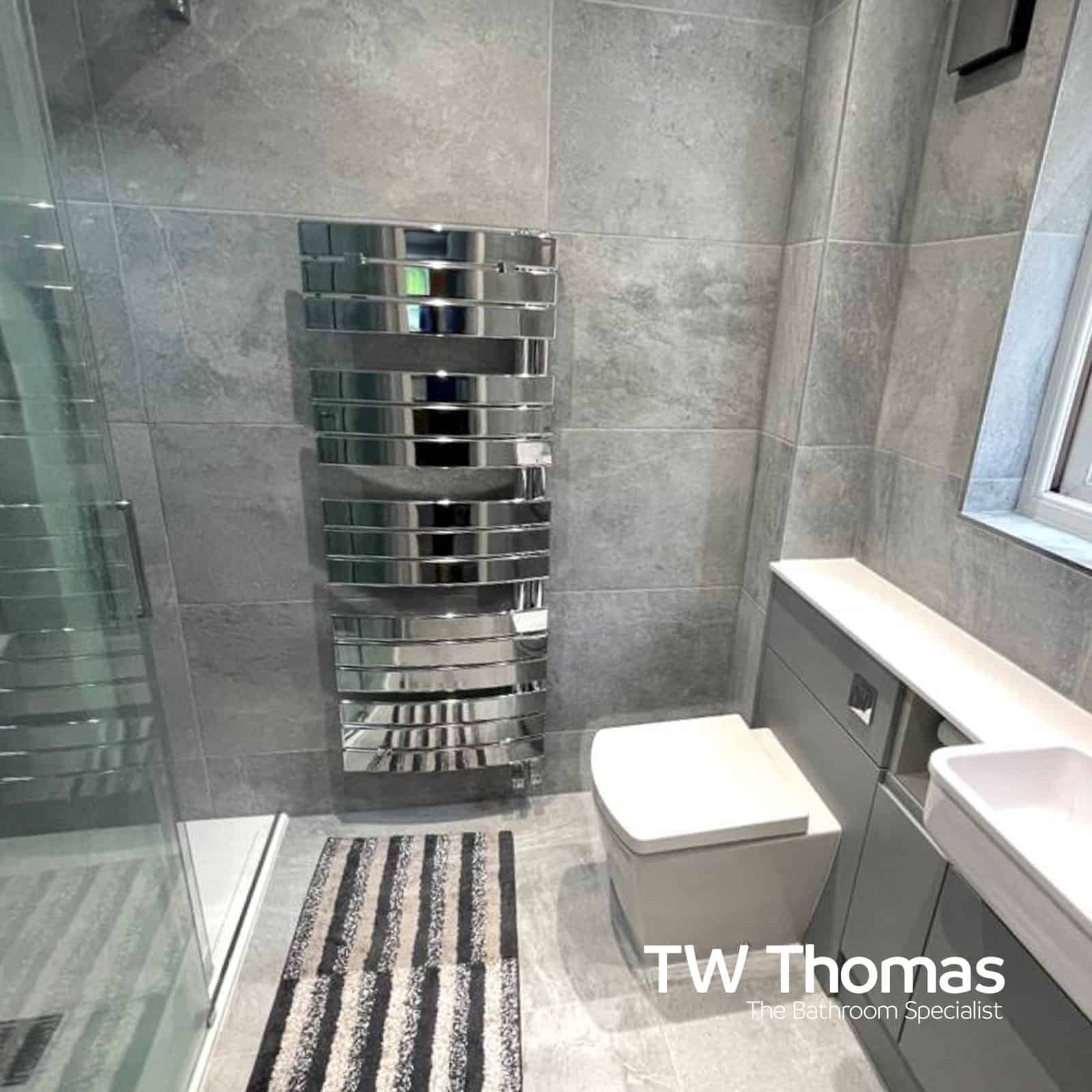 A grey tiled shower room with a large chrome designer towel radiator, a wall-hung WC and a glass shower screen