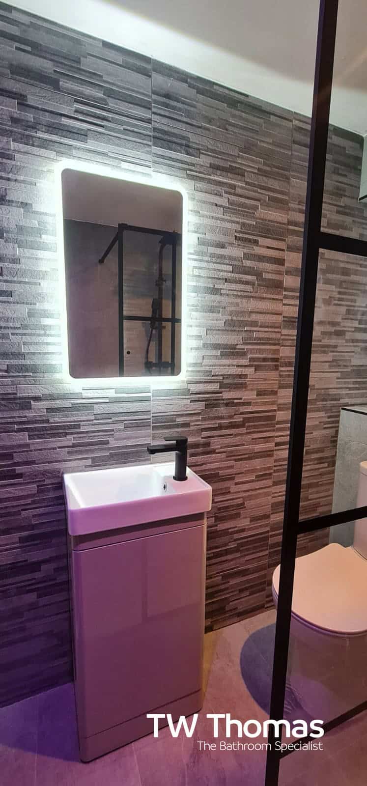 A pink gloss cloakroom unit with a white basin and matt black tap below a backlit mirror, against a brick-effect mosaic wall