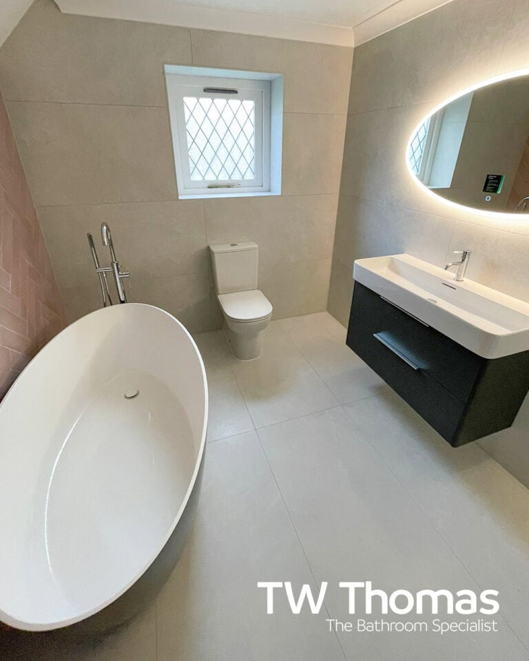 A white oval freestanding bath with a chrome floor-standing filler, a back-to-wall WC under a leaded window and a large oval backlit mirror