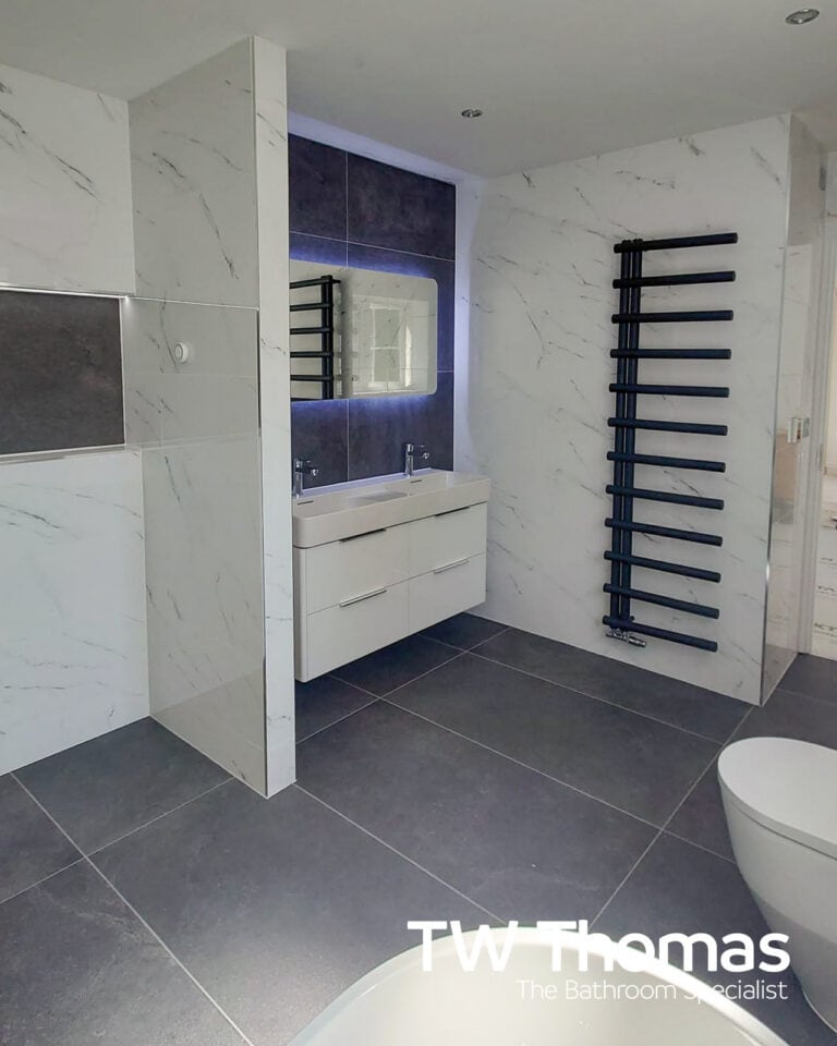 Twin basins on a white wall-hung double vanity against marble-effect wall tiles, with a backlit mirror and a black ladder towel radiator