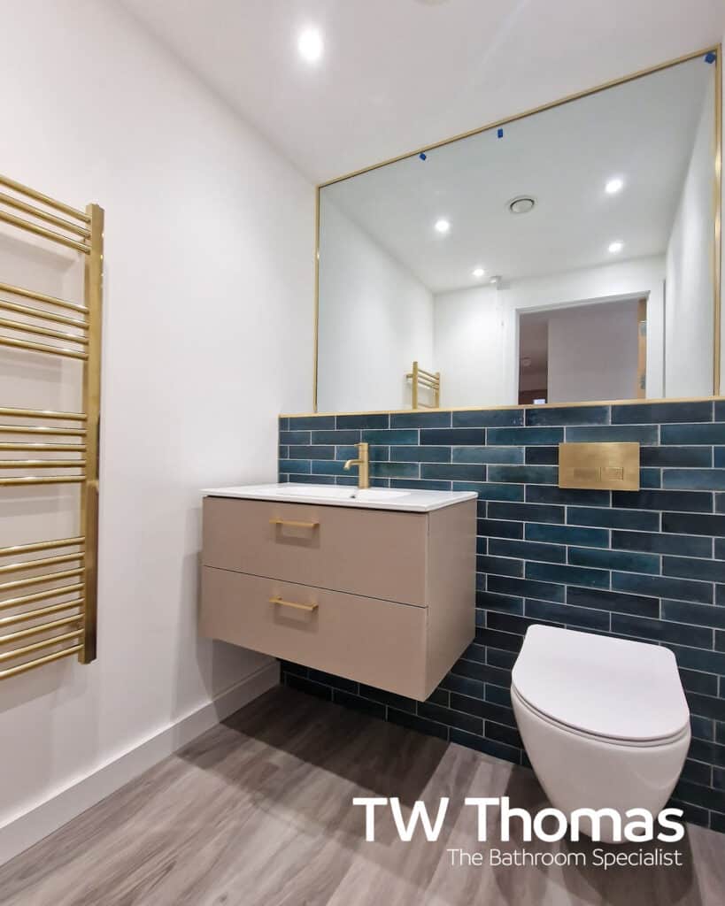 A wall-hung WC with a brushed brass flush plate against dark blue brick-effect tiles, beside a taupe wall-hung vanity and a brass framed mirror