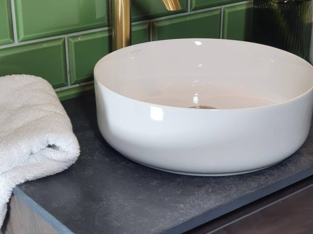 Armera round ceramic countertop bowl on a dark stone top with a brushed gold tap against green metro tiles