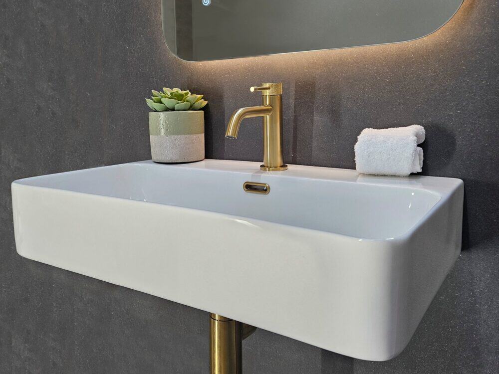 Armera Reef wall-hung ceramic basin with a brushed gold mixer below a backlit mirror