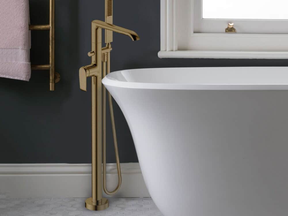 Armera Vaere floor-standing bath shower mixer in brushed gold beside a freestanding bath and a brushed gold towel warmer