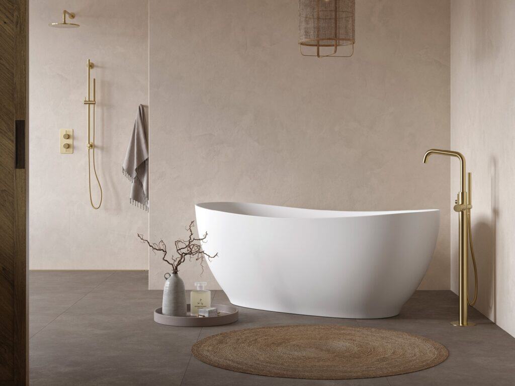 Adamsez curved freestanding bath in a warm plaster bathroom with a brushed brass floor-standing mixer and a round jute rug