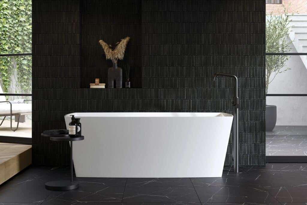 Adamsez freestanding bath against a black tiled wall with a matt black floor-standing mixer and a garden beyond