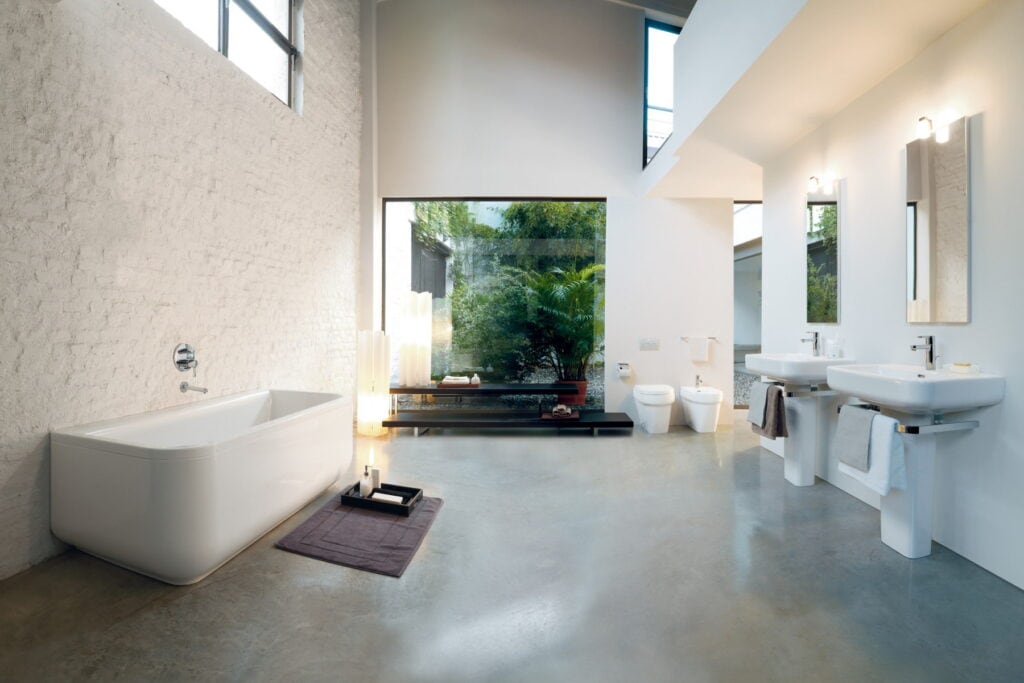 Laufen bathroom in a white-brick loft with a bath, a back-to-wall WC and bidet and two pedestal washbasins below wall mirrors