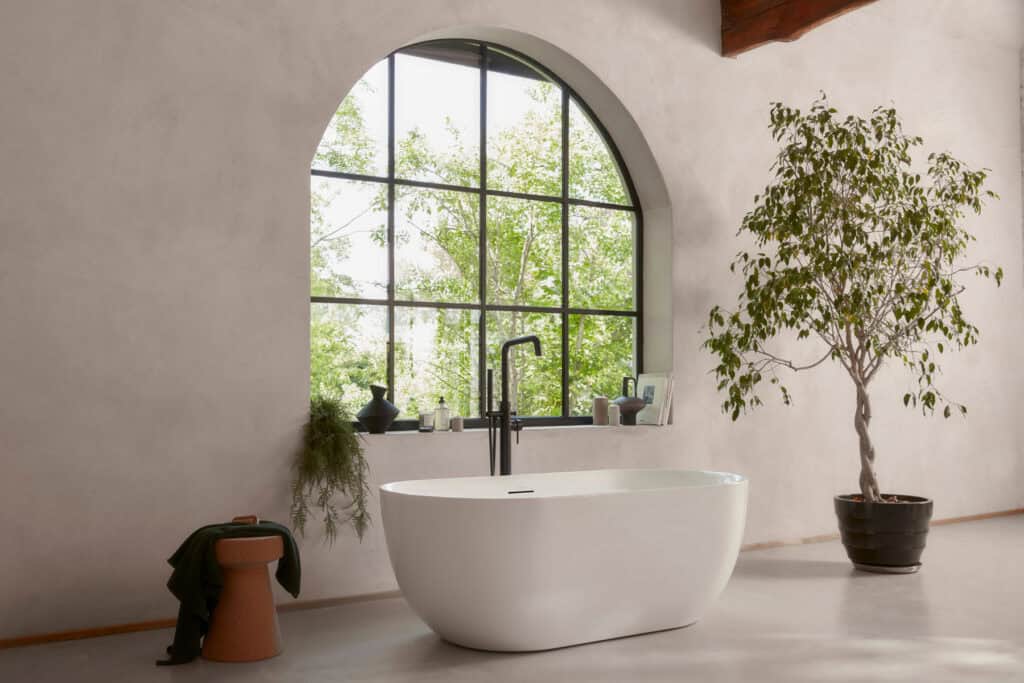Roper Rhodes Note freestanding bath beneath an arched window, with an olive tree and plaster walls