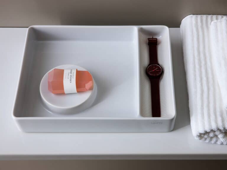 Laufen VAL white ceramic tray holding a soap dish and a watch, beside rolled towels