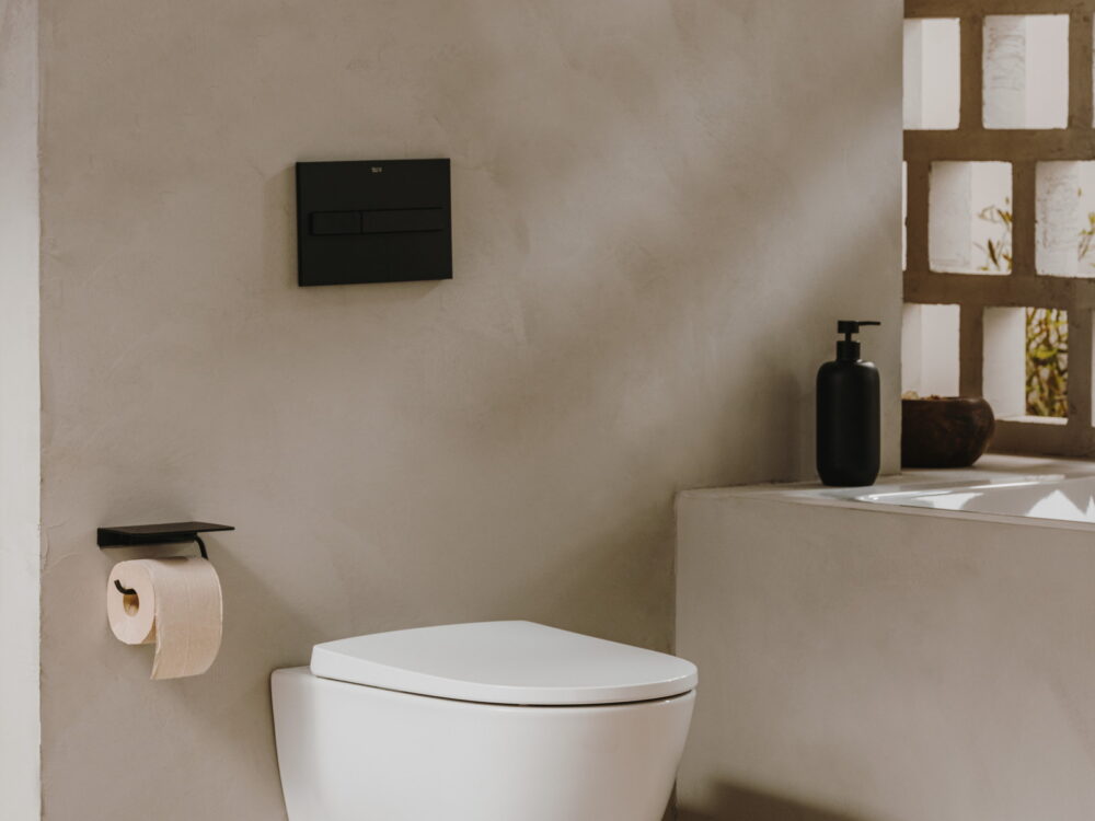 Roca Ona wall-hung WC with a matt black flush plate and paper holder in a plaster-walled cloakroom