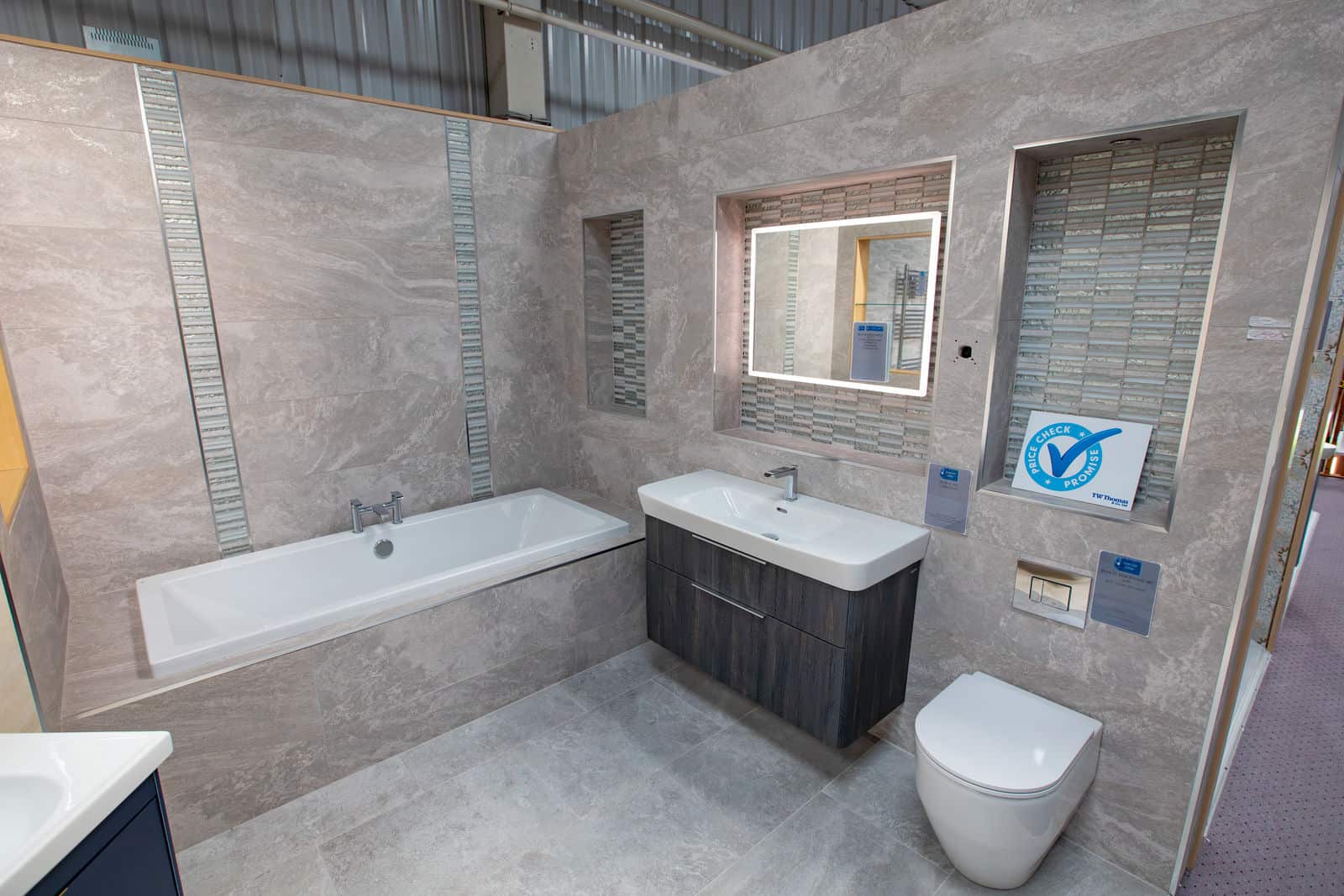 A bath, grey vanity unit and WC in a grey tiled room with an illuminated mirror